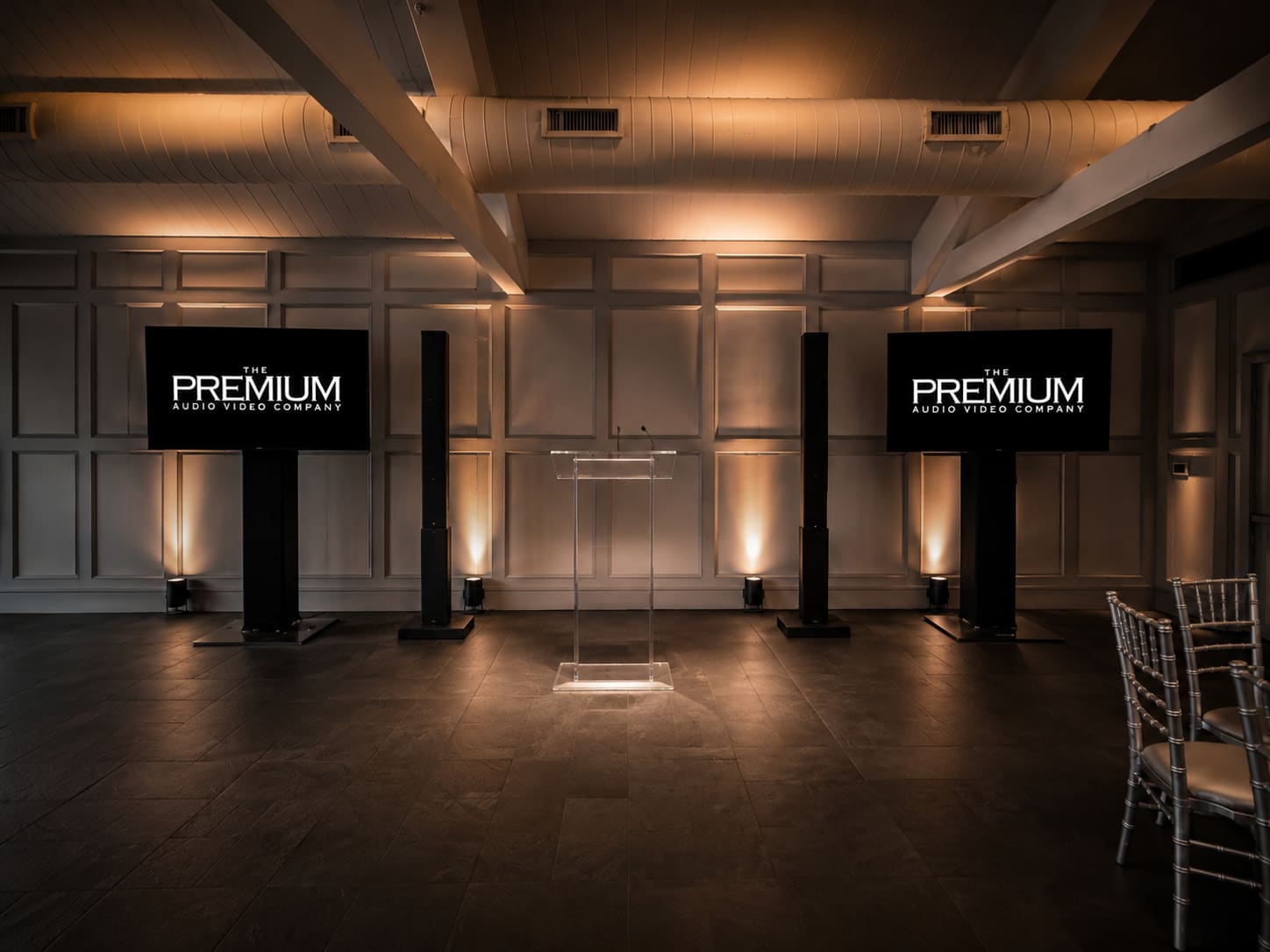 Ballroom event setup with two PremiumAVC-branded flat screens flanking an acrylic lectern, warm amber uplighting, and Chiavari seating